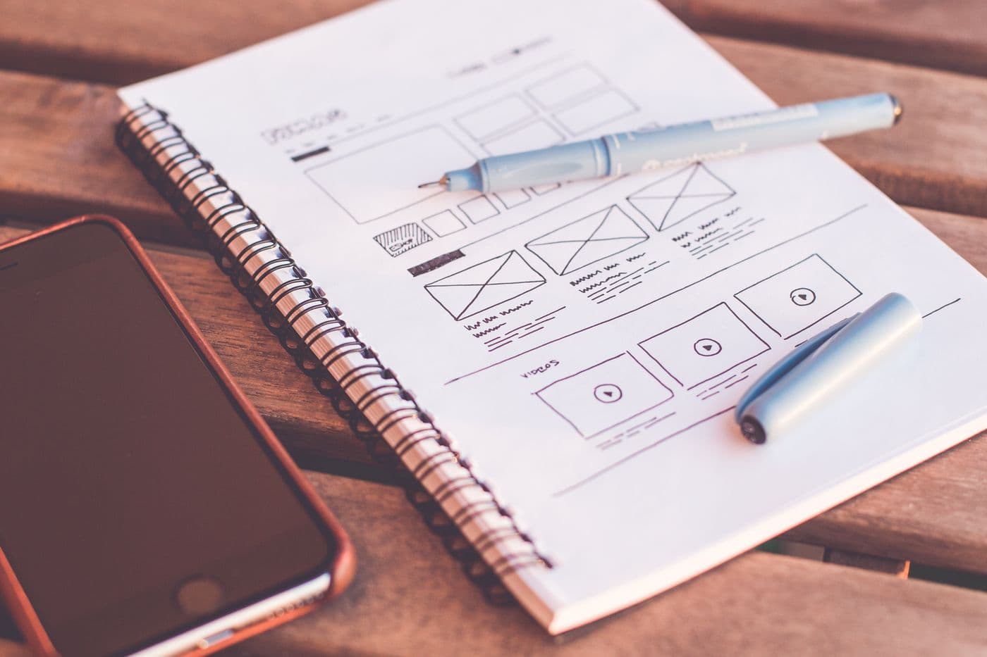 Sketsa wireframe desain di notebook spiral dengan pena dan ponsel di atas meja kayu