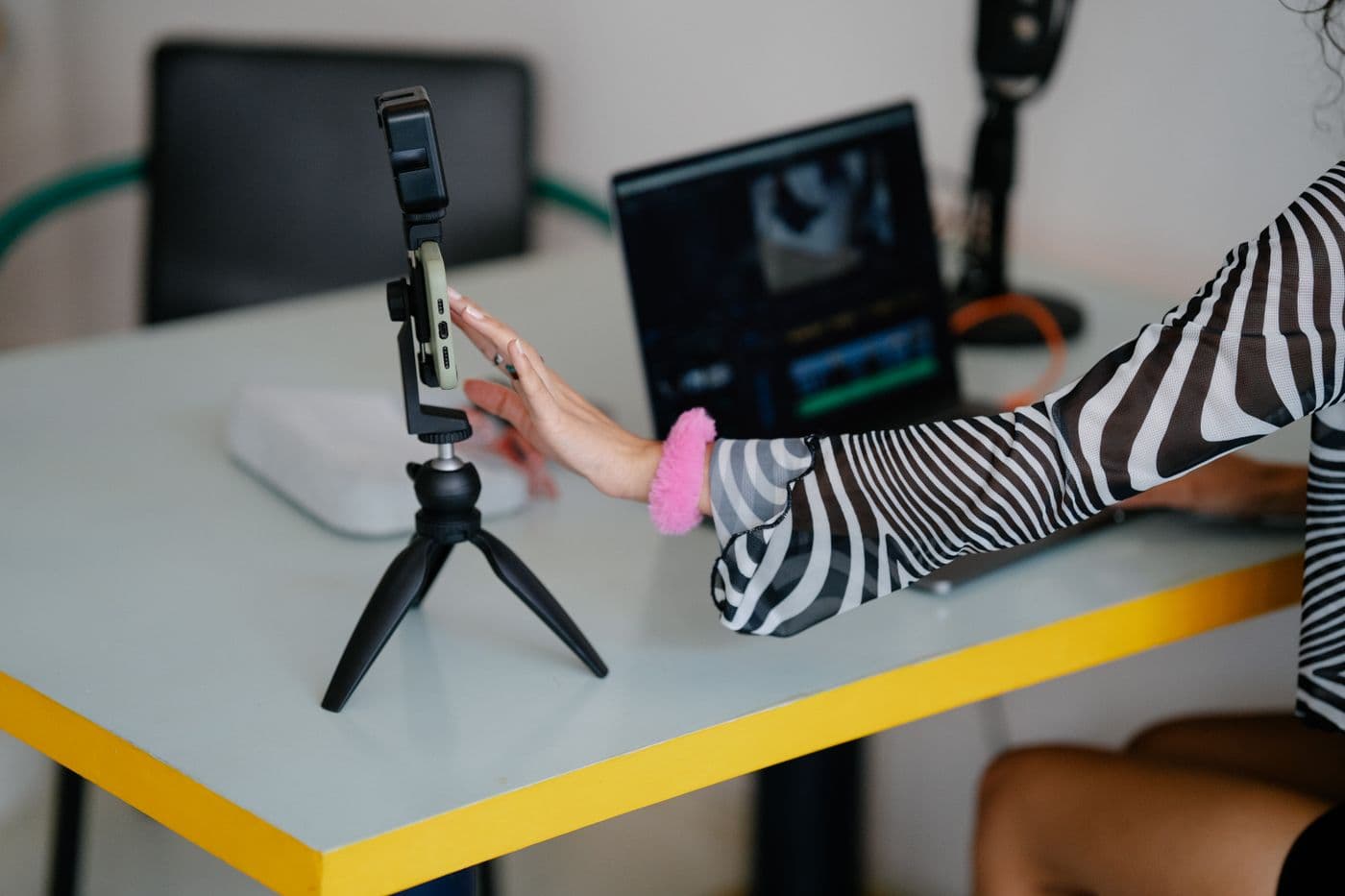 Ponsel di atas tripod mini di samping laptop yang menampilkan timeline penyuntingan video
