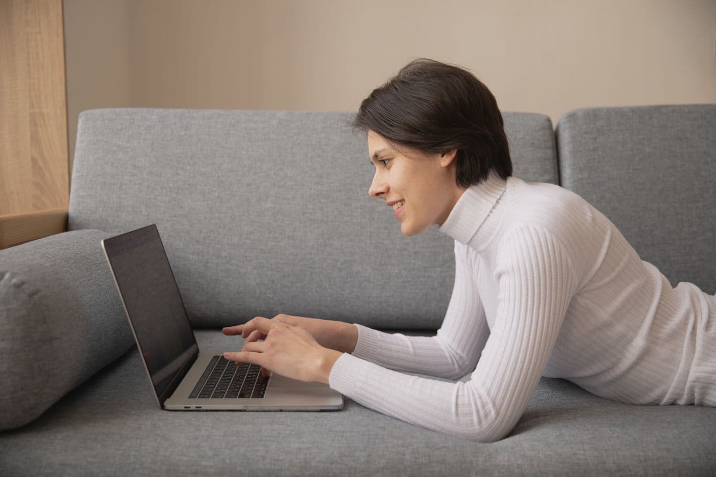 Perempuan berbaring di sofa abu-abu sambil tersenyum dan mengetik di laptop