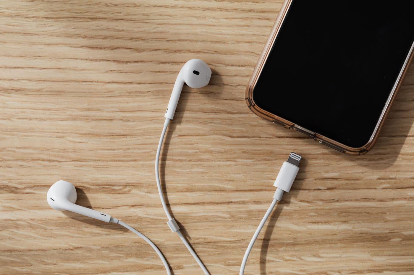 Earphone kabel putih tergeletak di samping ponsel berlayar hitam di atas meja kayu terang