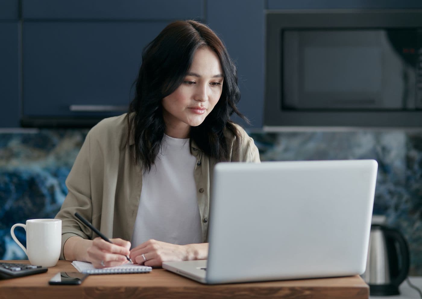 Perempuan mencatat di buku kecil sambil membaca layar laptop di meja kayu dengan cangkir putih di sampingnya