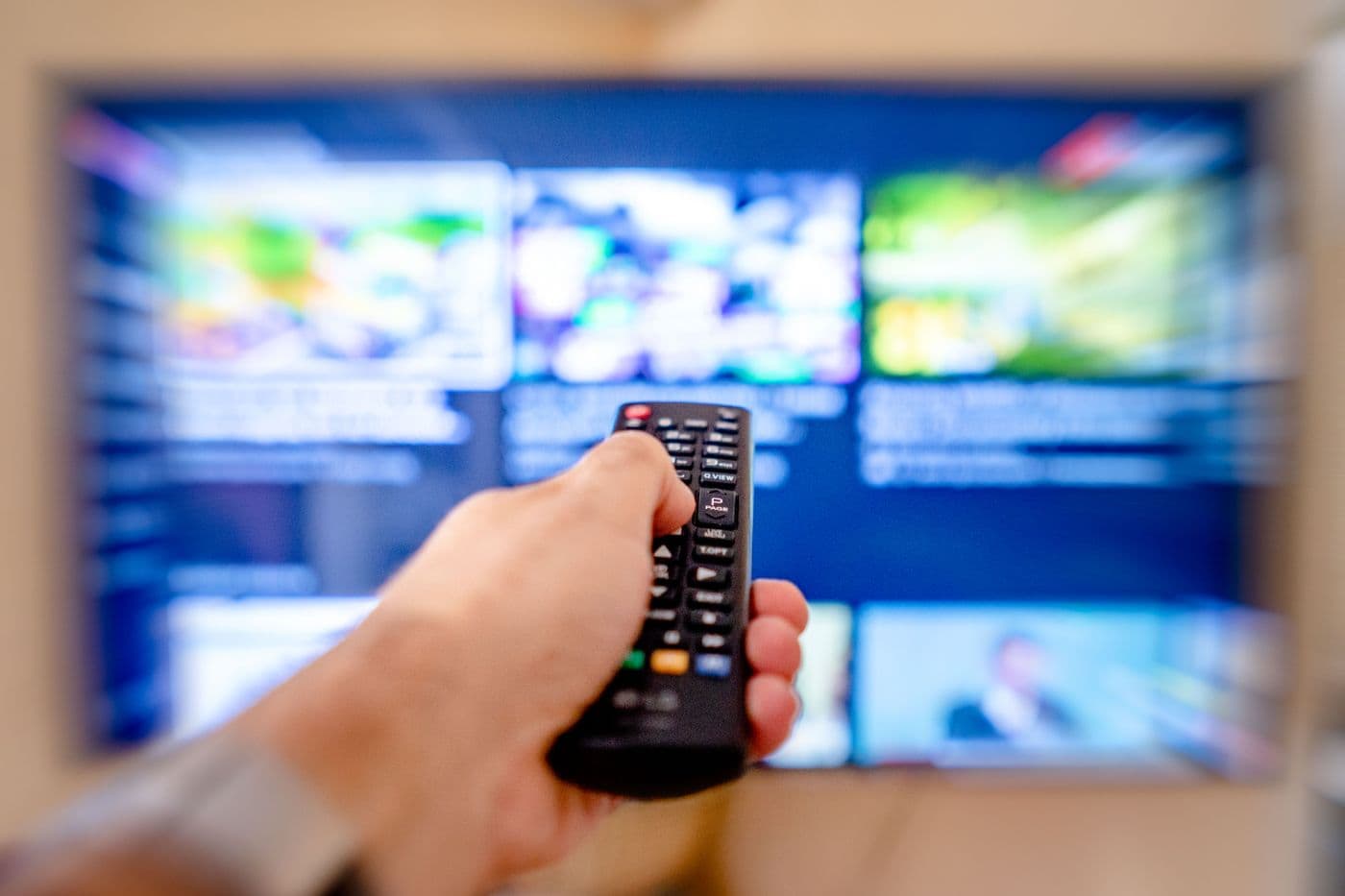 Tangan memegang remote TV yang diarahkan ke layar televisi berisi deretan tayangan yang tampak buram