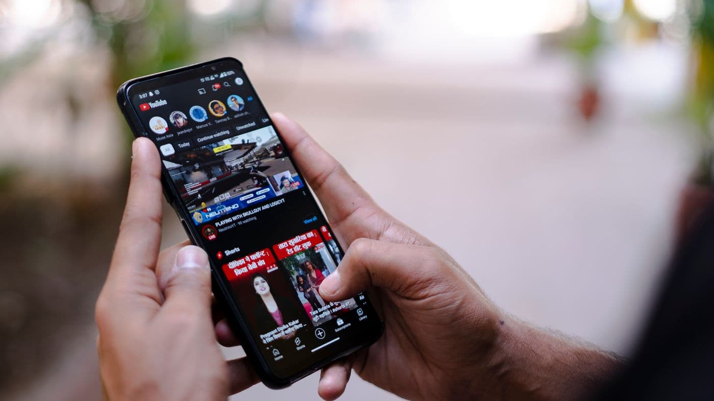 Dua tangan memegang ponsel yang menampilkan beranda aplikasi YouTube dengan latar luar ruangan yang buram