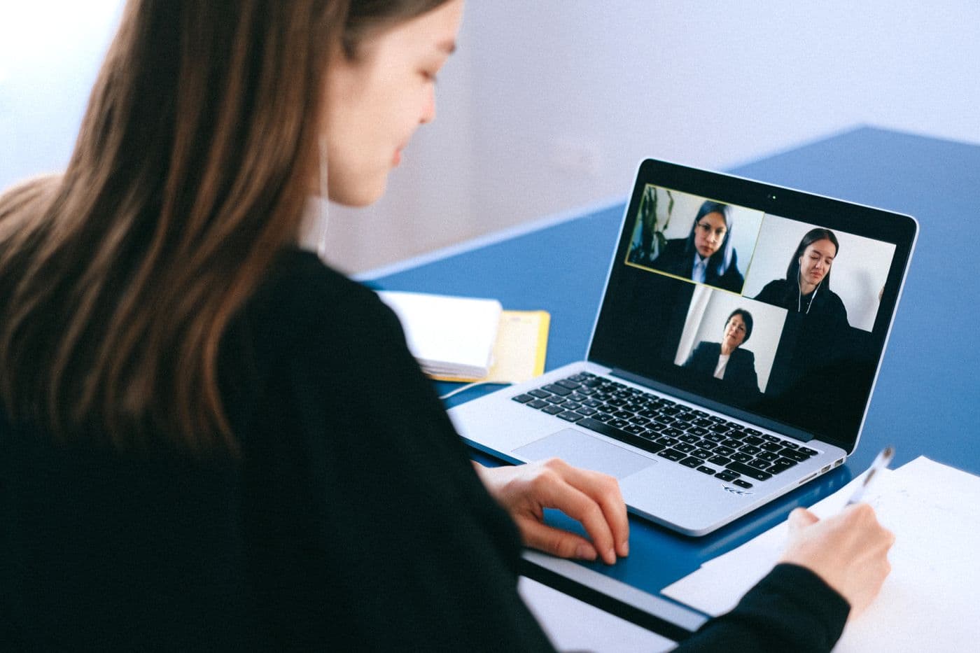 Perempuan mencatat di kertas sambil mengikuti rapat video di laptop yang menampilkan tiga peserta lain